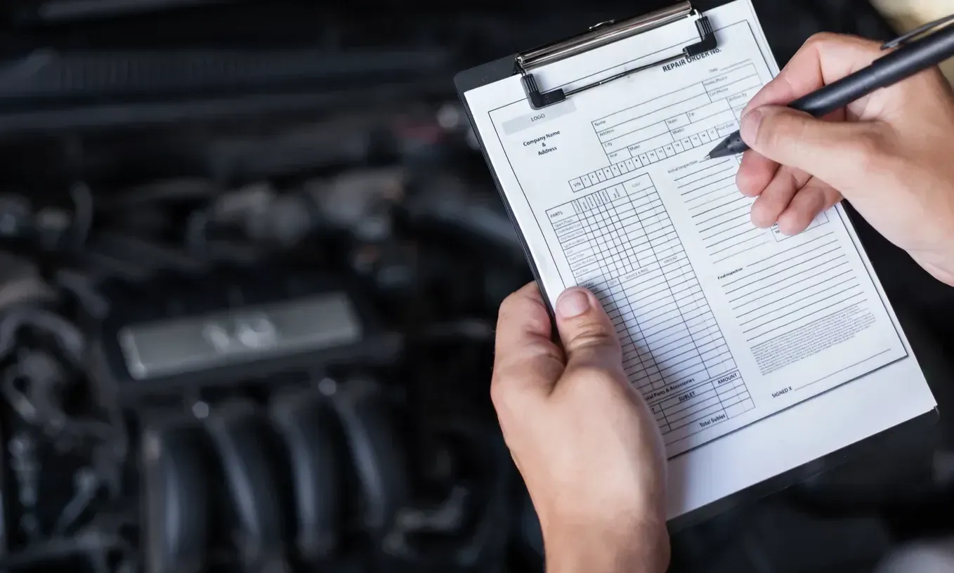 Manos sosteniendo documento y realizando anotación y de fondo un motor de auto