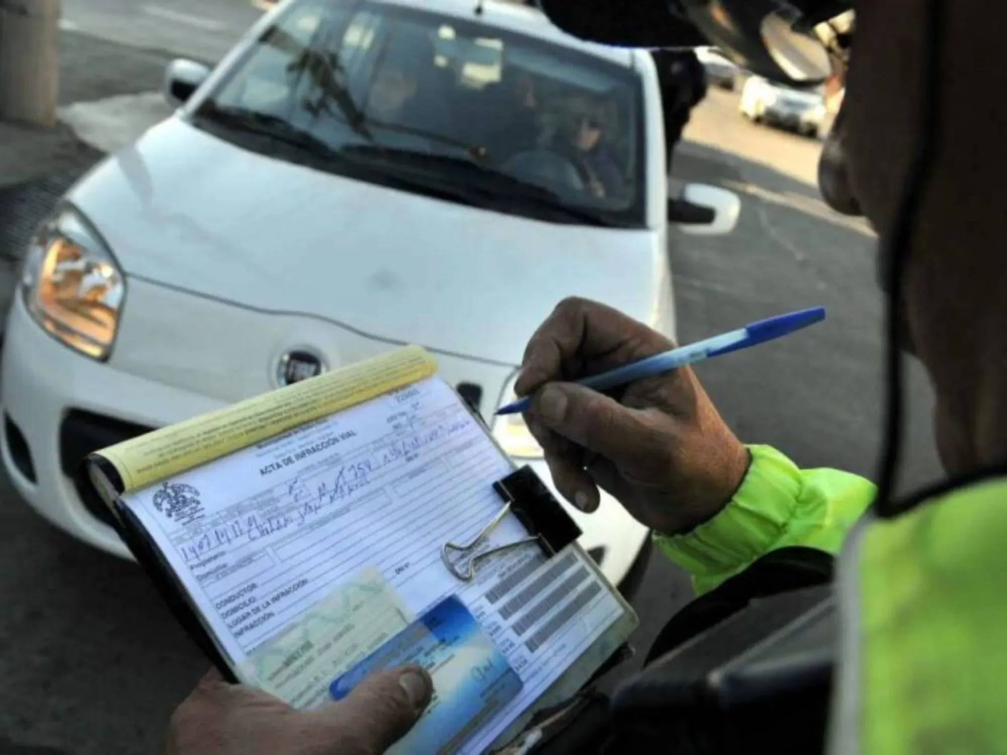 Persona de transito redactando multa y al fondo un auto blanco con personas abordo