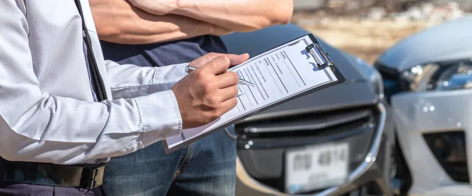 Persona sosteniendo documento y bolígrafo al lado de otra persona y al fondo dos autos