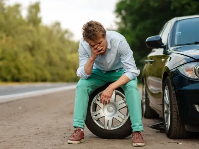 Persona con mano en la cara sobre neumático de auto y al lado un auto