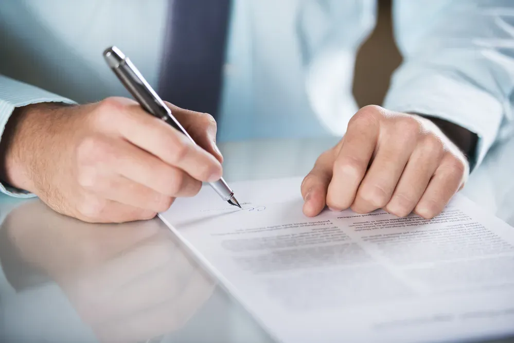 Mano sosteniendo un bolígrafo y firmando un documento sobre una mesa