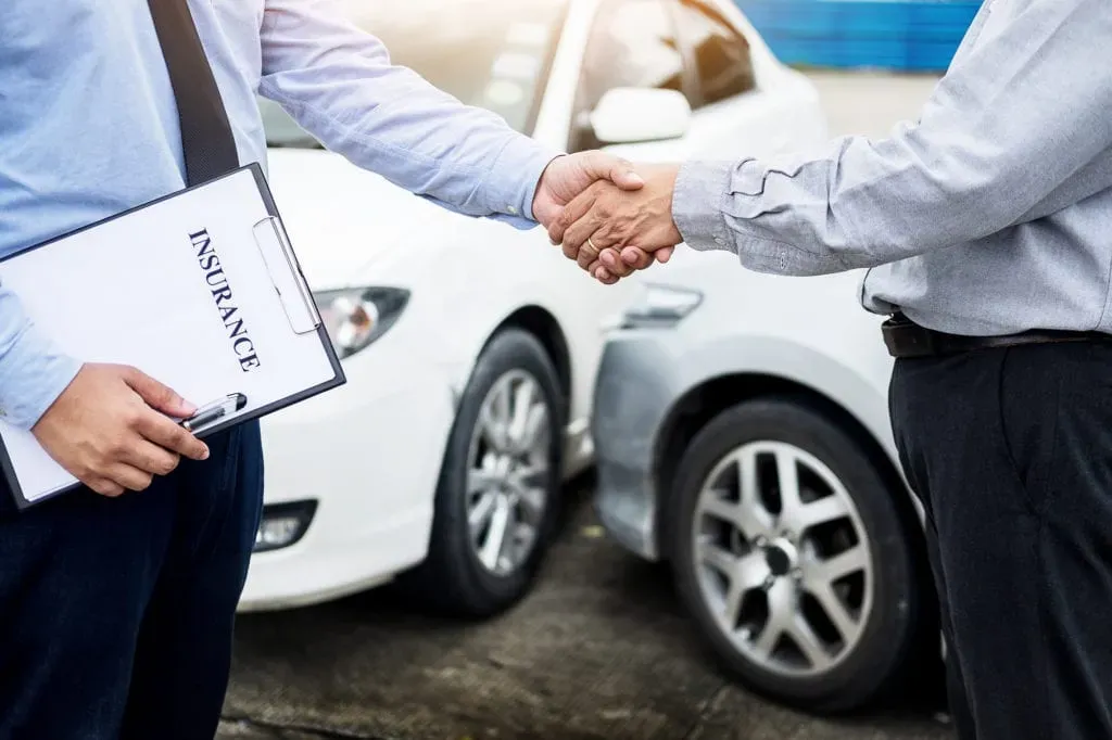 Personas estrechando manos sosteniendo un contrato y al fono autos color blanco muy cerca uno del otro