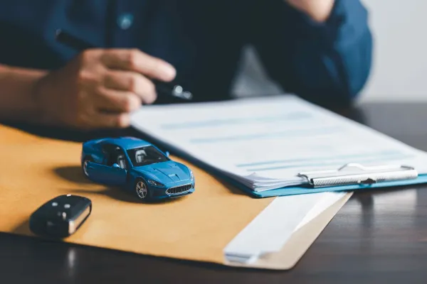 Sobre con papeles un auto azul de juguete con unas llaves de auto y al lado papeles y una mano con bolígrafo