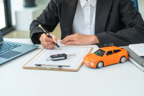 Mano sosteniendo un boligrafo sobre un contrato en una mesa con una llave de autos y un carro de juguete naranja