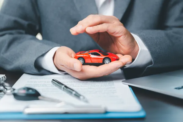 Manos alrededor de un mini auto rojo de colección y un contrado con una llave de auto y pluma