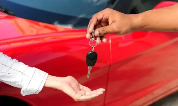 Mano sosteniendo una llave de coche con otra mano recibiendola y un auto rojo de fondo