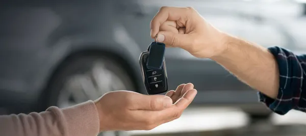 Mano sosteniendo llaves de auto y otra mano recibiendola y un auto de fondo