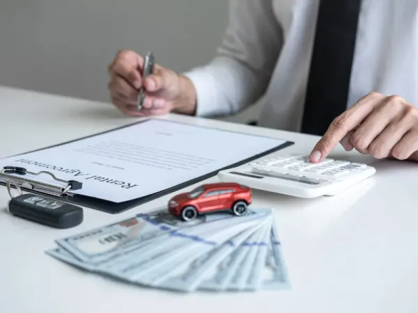 Persona tocando calculadora  y sosteniendo un bolígrafo sobre una mesa con un documento dinero y un auto a escala rojo