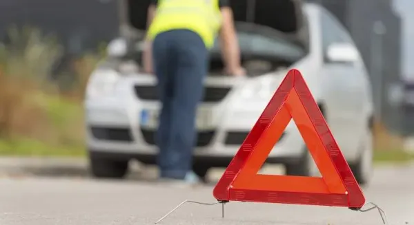 Triangulo de emergencia para carretera y al fondo auto con capo levantado y persona con chaleco amarillo revisándolo 