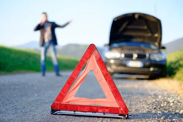 Triangulo de emergencia colocado en carretera y al fondo un auto con capo levantado y una persona con los brazos abiertos